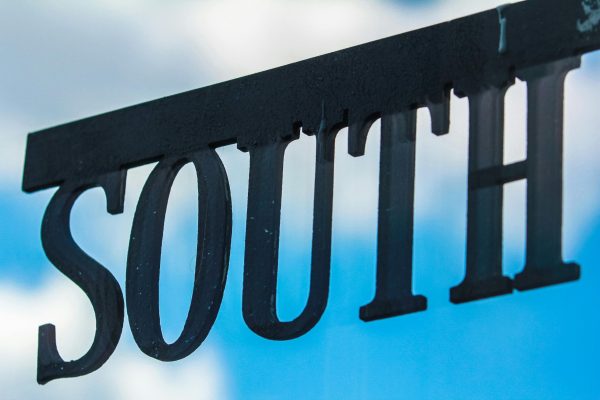 Minimalist metal 'south' sign silhouetted against a bright blue sky with cloud background.
